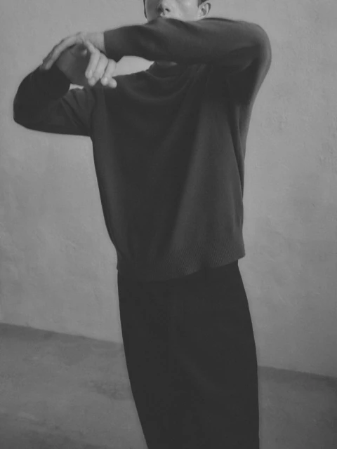 Monochrome image of an adult posing in a sweater and pants, with a soft, blurred quality, set against a mottled grey backdrop.