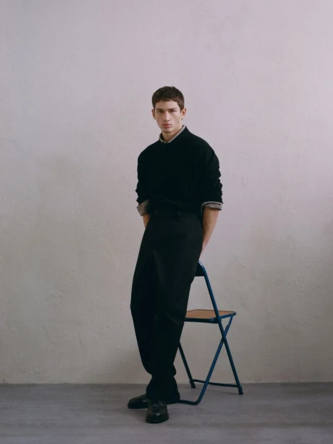 Man in a black sweater and trousers poses indoors, leaning against a blue folding chair in a fashion editorial shot.