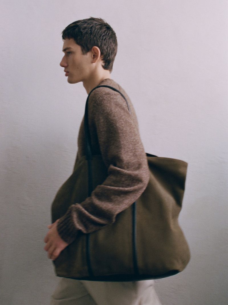 A young adult male in a brown knit sweater carries an olive green tote bag in a minimalist indoor setting with soft, diffused lighting.