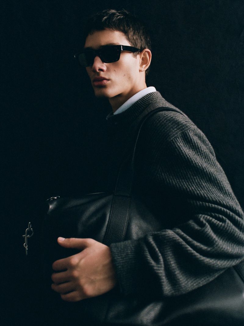 Model wearing dark sunglasses and a grey knit sweater holding a black leather bag against a dark background for Massimo Dutti Punto.