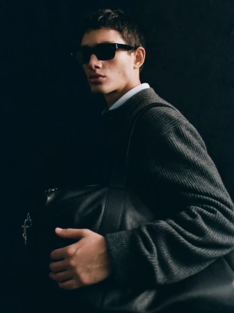 Model wearing dark sunglasses and a grey knit sweater holding a black leather bag against a dark background for Massimo Dutti Punto.