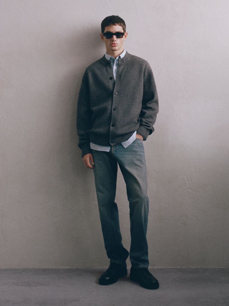 Full-length shot of an adult male wearing a grey cardigan, light blue shirt, jeans, sunglasses, and black shoes, standing in a neutral indoor space.
