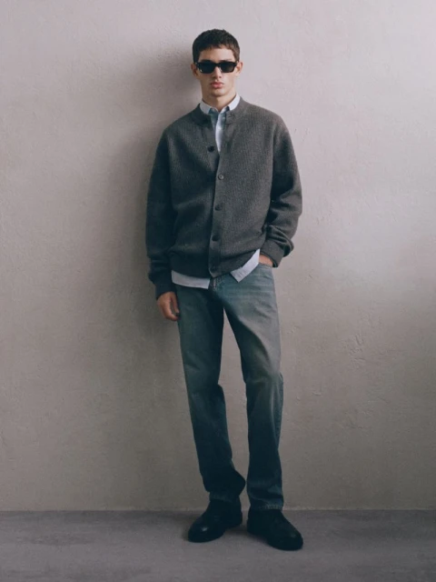 Full-length shot of an adult male wearing a grey cardigan, light blue shirt, jeans, sunglasses, and black shoes, standing in a neutral indoor space.