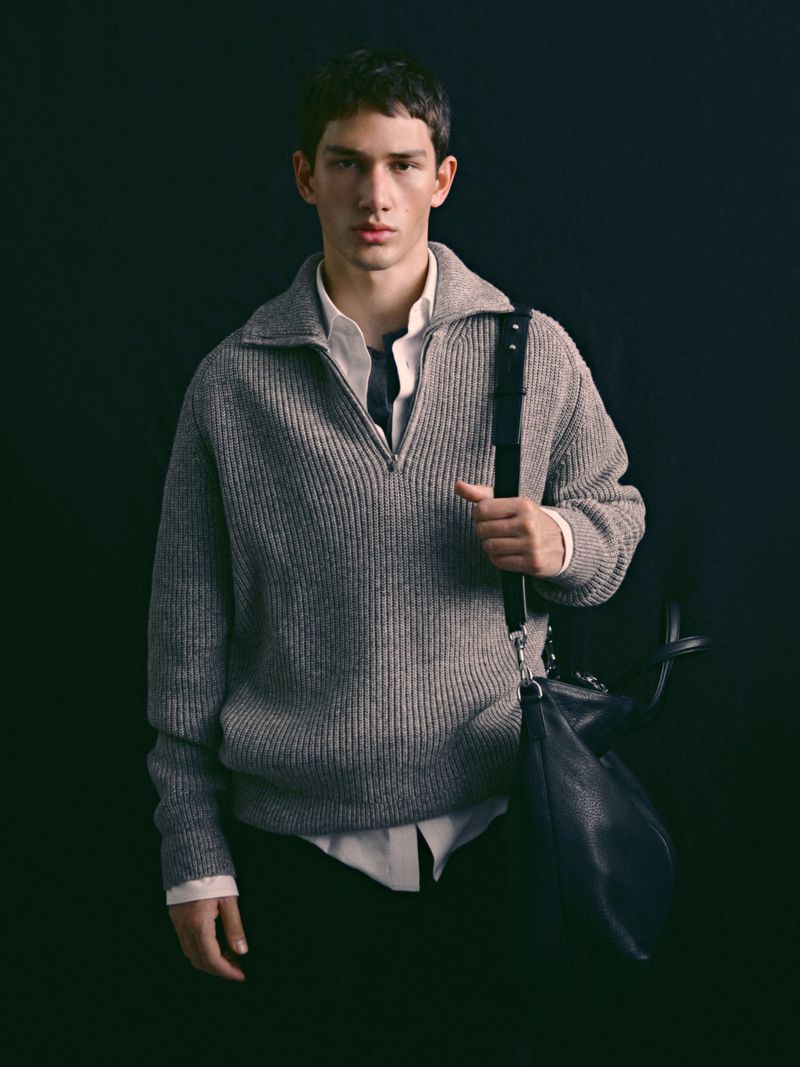 Portrait of a young adult male wearing a gray zip-up sweater and carrying a black bag with shoulder strap against a dark background.