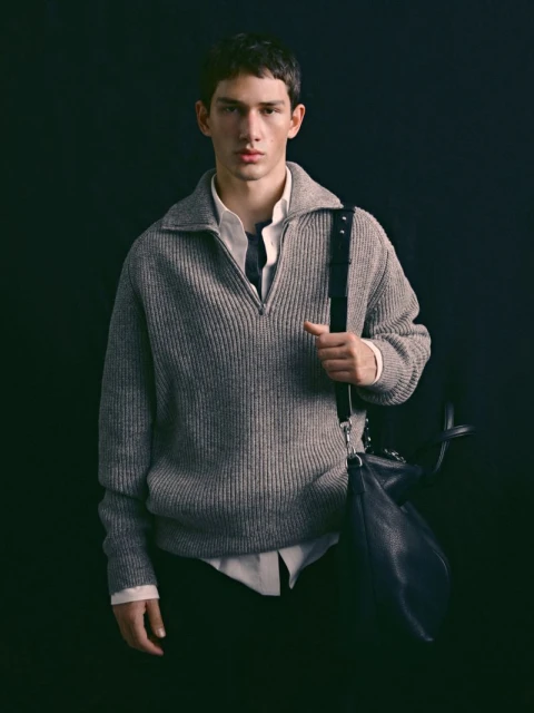 Portrait of a young adult male wearing a gray zip-up sweater and carrying a black bag with shoulder strap against a dark background.