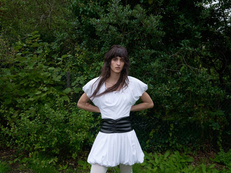 An adult woman in a white dress with a black belt poses outdoors in a lush green environment. She looks directly at the camera.