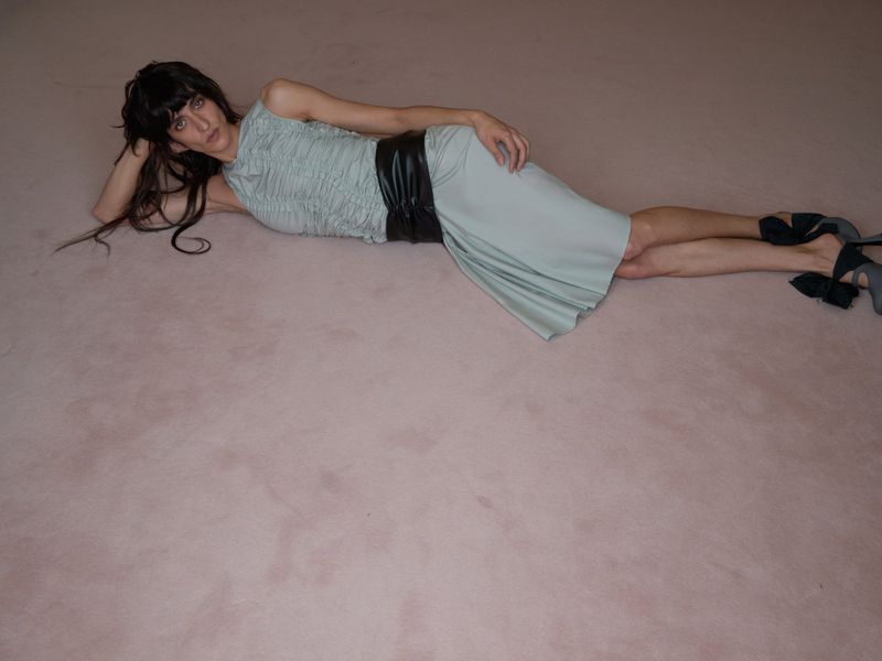 A woman in a light blue dress lies on a pink surface, wearing black heels and a black belt; shot at eye-level with a full shot.