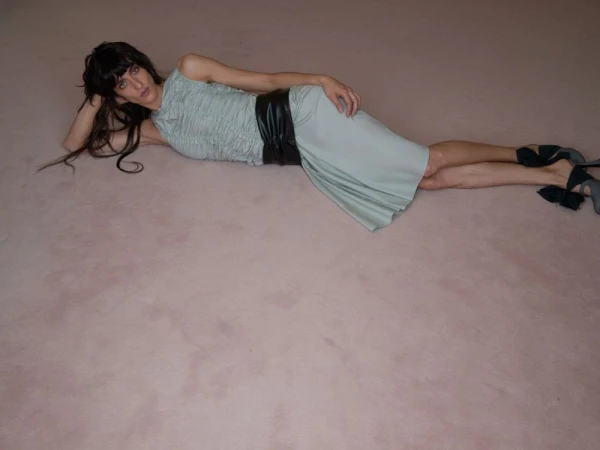 A woman in a light blue dress lies on a pink surface, wearing black heels and a black belt; shot at eye-level with a full shot.