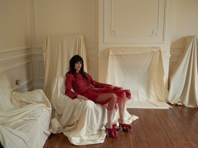 Adult woman in a red dress seated on a covered chair in an off-white room, wearing red shoes with bows and posing for a fashion shot.
