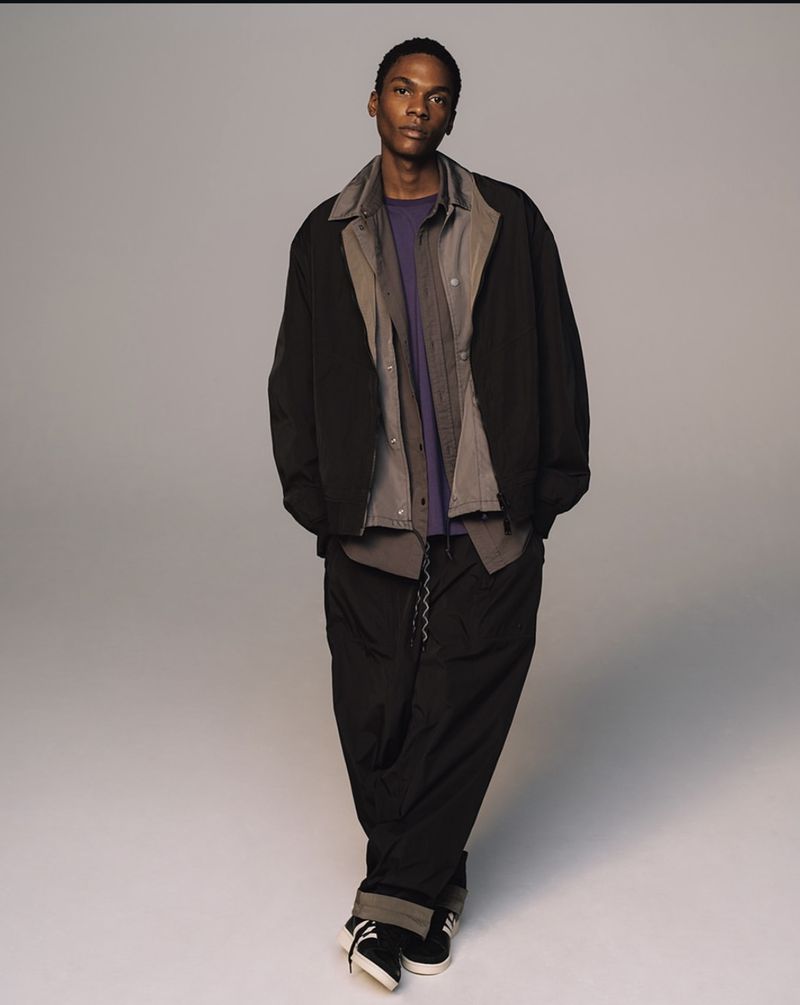 A full-length studio shot of a slender adult male with dark skin wearing a jacket, collared shirt, t-shirt, pants, and sneakers.