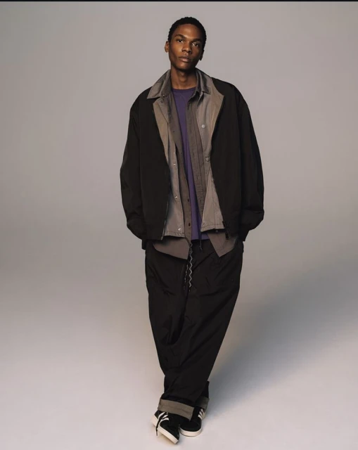 A full-length studio shot of a slender adult male with dark skin wearing a jacket, collared shirt, t-shirt, pants, and sneakers.
