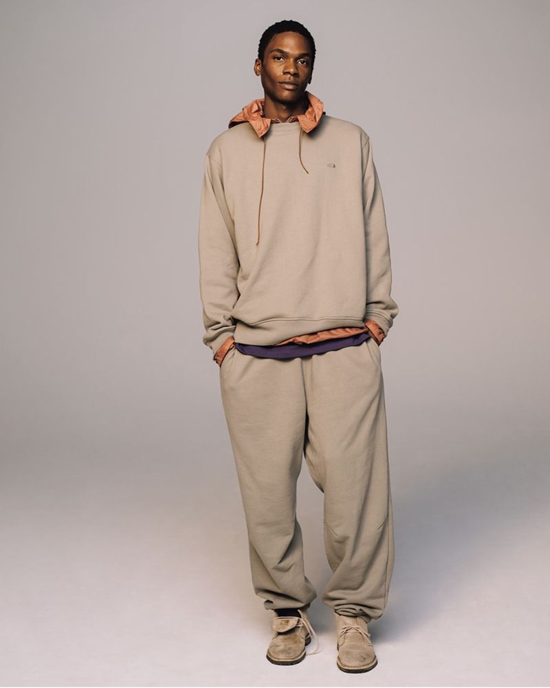An adult male poses in a beige hooded sweatshirt and matching sweatpants in a full-body shot against a neutral grey background.