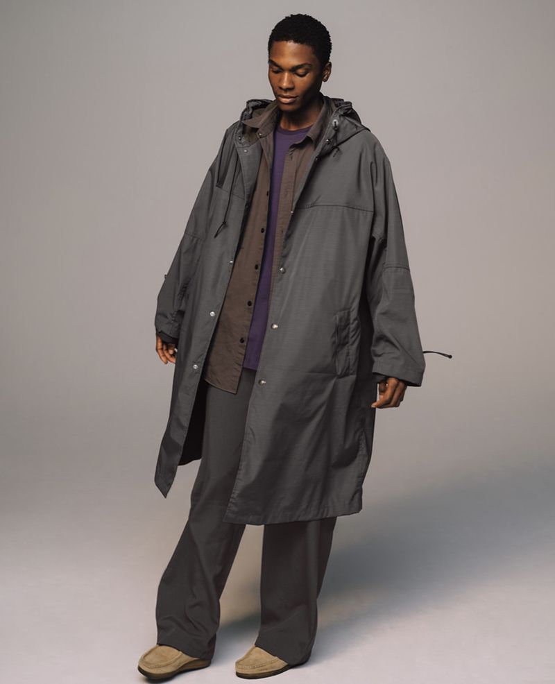A Black male model poses in a grey hooded raincoat, purple shirt, and grey trousers in a photo studio with soft lighting.