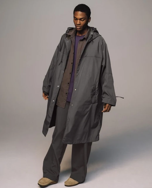 A Black male model poses in a grey hooded raincoat, purple shirt, and grey trousers in a photo studio with soft lighting.