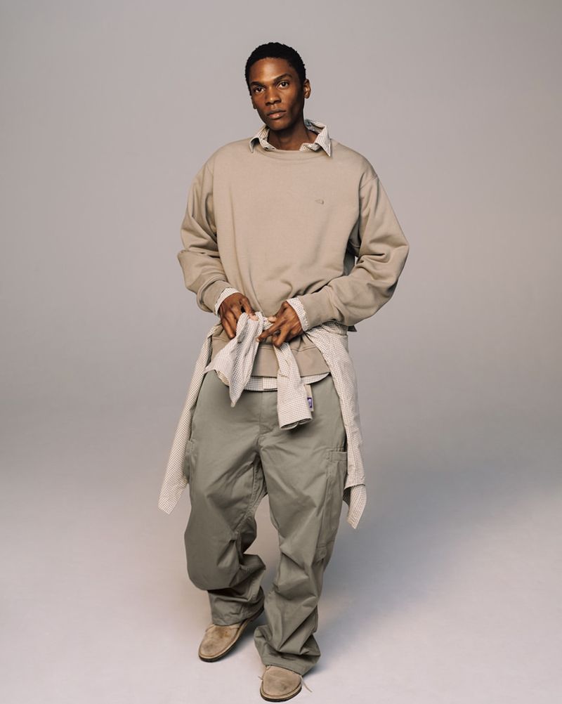 Studio shot of a Black male fashion model in khaki cargo pants, a beige sweatshirt, and suede shoes, standing against a gray backdrop.