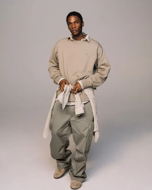 Studio shot of a Black male fashion model in khaki cargo pants, a beige sweatshirt, and suede shoes, standing against a gray backdrop.