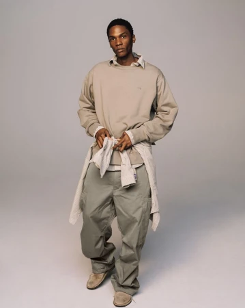 Studio shot of a Black male fashion model in khaki cargo pants, a beige sweatshirt, and suede shoes, standing against a gray backdrop.