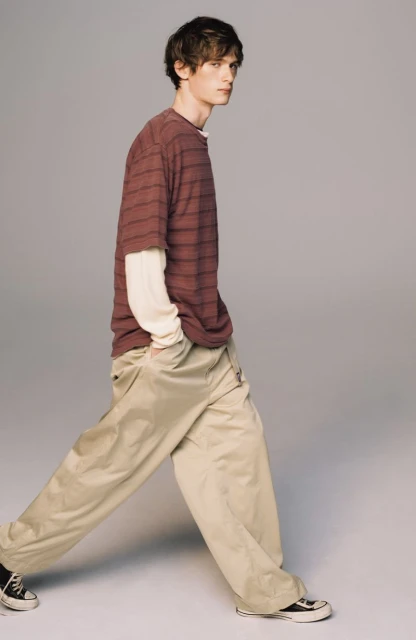 Full-length shot of a young adult male walking in baggy beige pants and a striped t-shirt against a neutral grey backdrop.