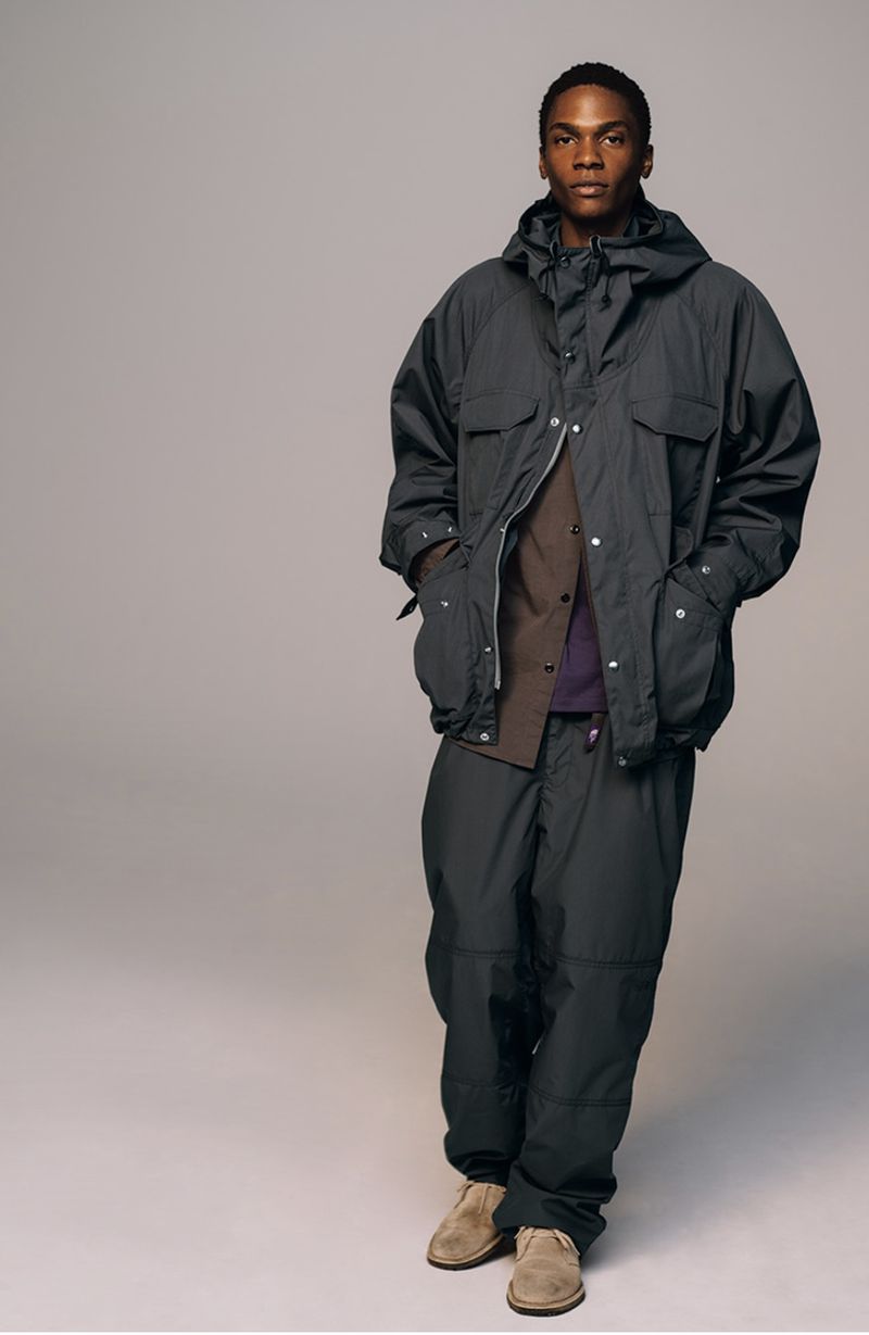 A man in a dark grey hooded jacket and pants stands indoors against a grey background in a studio shot, looking at the camera.