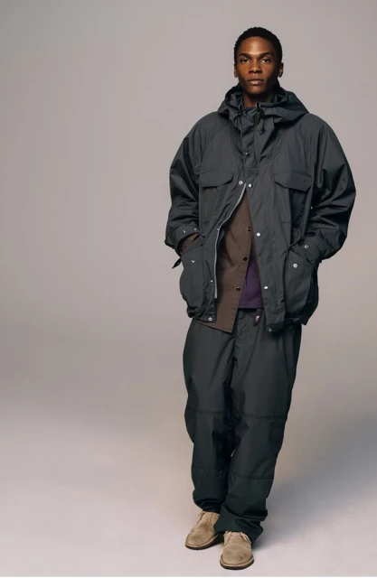 A man in a dark grey hooded jacket and pants stands indoors against a grey background in a studio shot, looking at the camera.