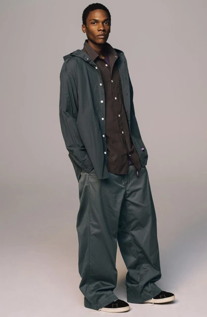 Full body studio shot of a young Black man in a green hooded jacket, brown button-down shirt, oversized pants, and sneakers against a neutral background.