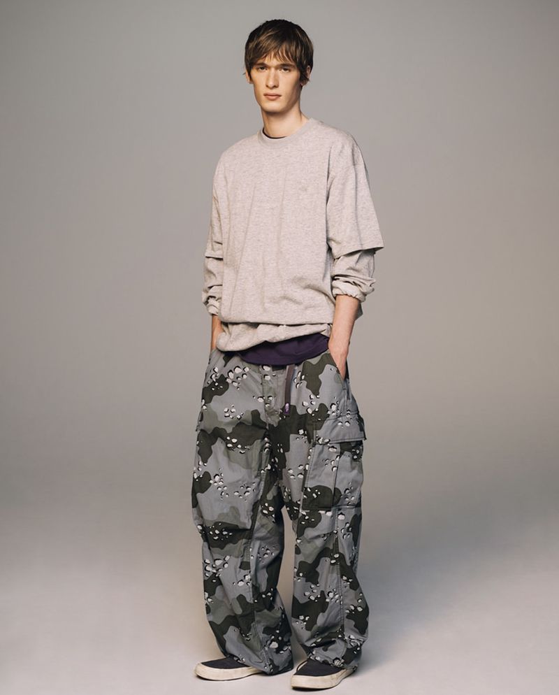 Full-shot of a young adult male in a grey long-sleeve shirt and camouflage pants standing in front of a neutral grey background.