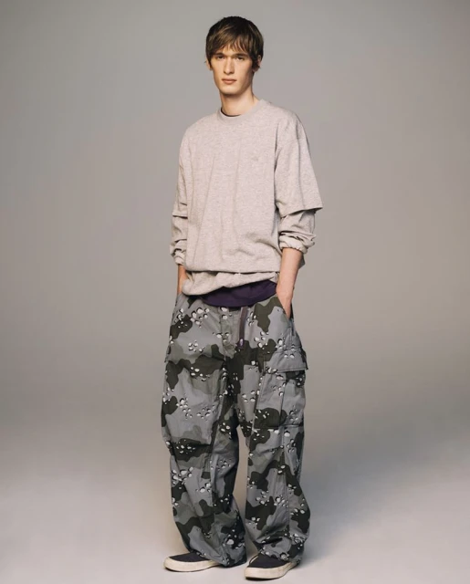Full-shot of a young adult male in a grey long-sleeve shirt and camouflage pants standing in front of a neutral grey background.