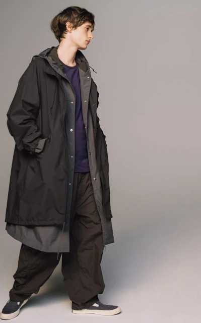 An adult male model poses in a hooded black raincoat, purple tee shirt, and dark trousers against a neutral gray studio backdrop. Fashion editorial.