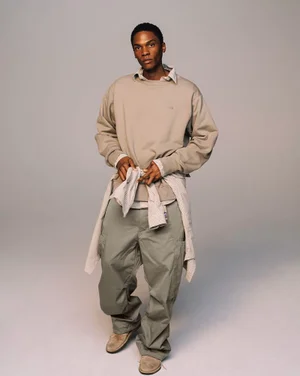 Model wearing a beige crewneck sweatshirt layered with a button-down shirt and light green cargo pants against a plain grey studio backdrop.