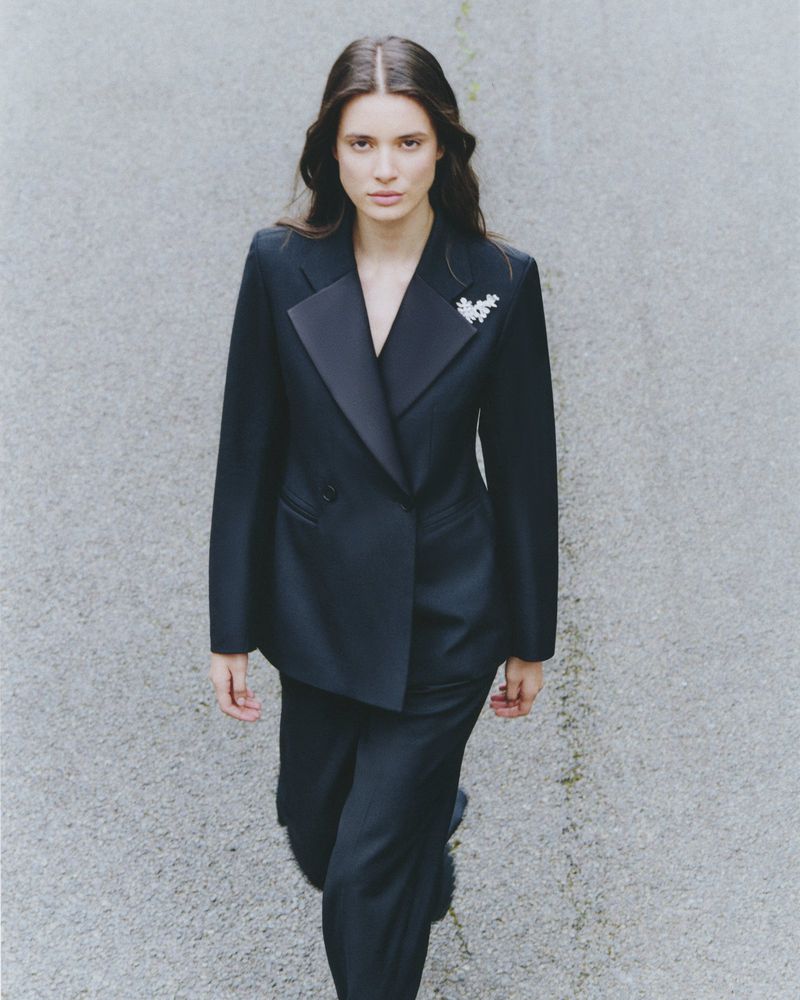 A woman in a black double-breasted suit walks on a concrete surface. She has long dark hair and wears a brooch on her lapel.