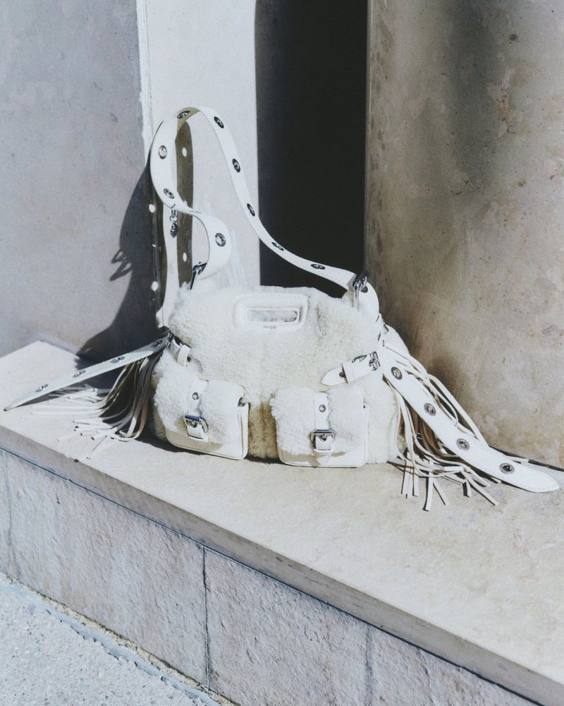 White fringed bag with white strap and silver hardware displayed indoors, near a gray textured pillar, fashion accessory advertisement.