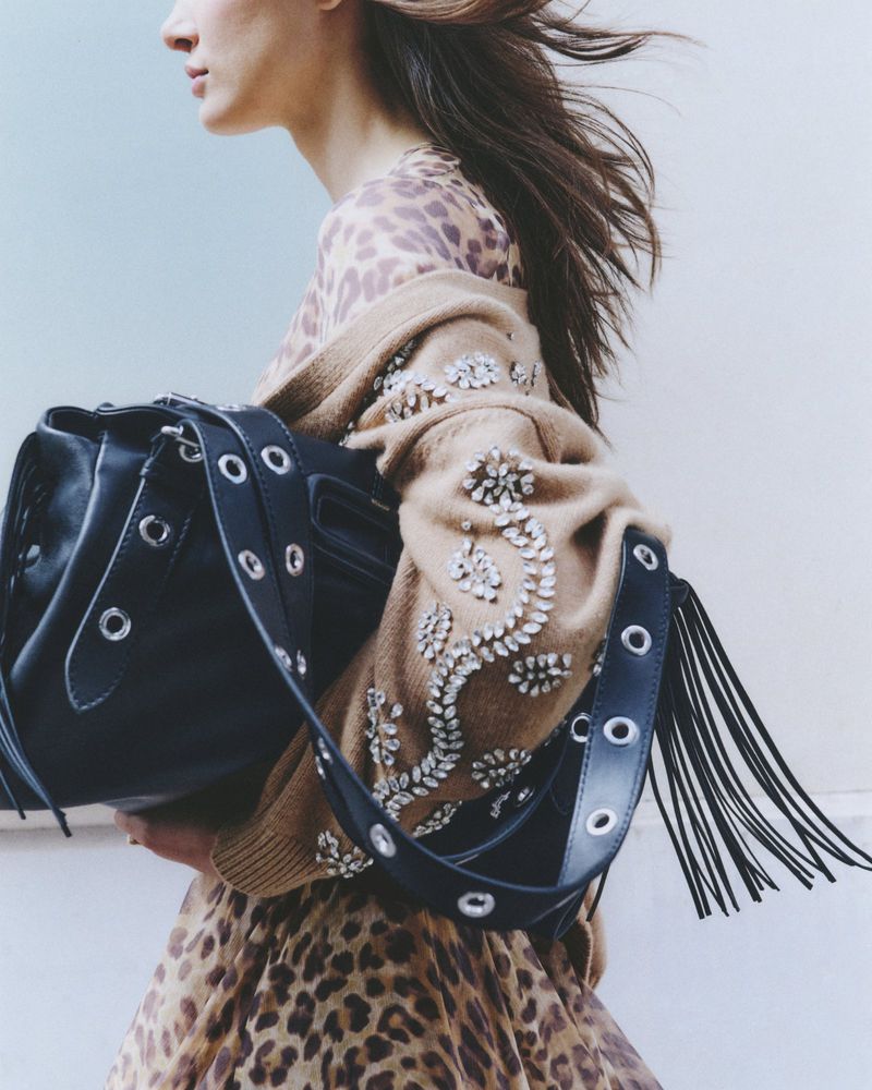 Woman wearing a leopard print dress and a tan, embellished cardigan with a black leather purse and fringed strap over her shoulder.