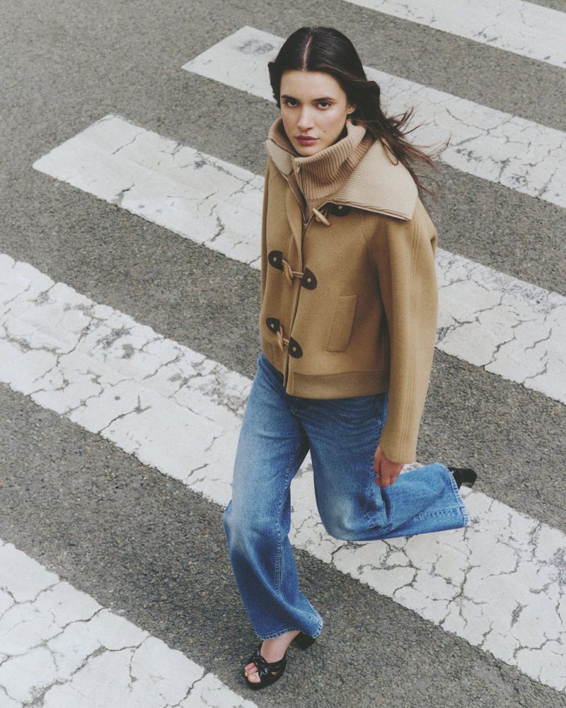 Woman in a tan duffle coat and jeans walks across a crosswalk with white stripes. Looking at camera from an overhead angle.
