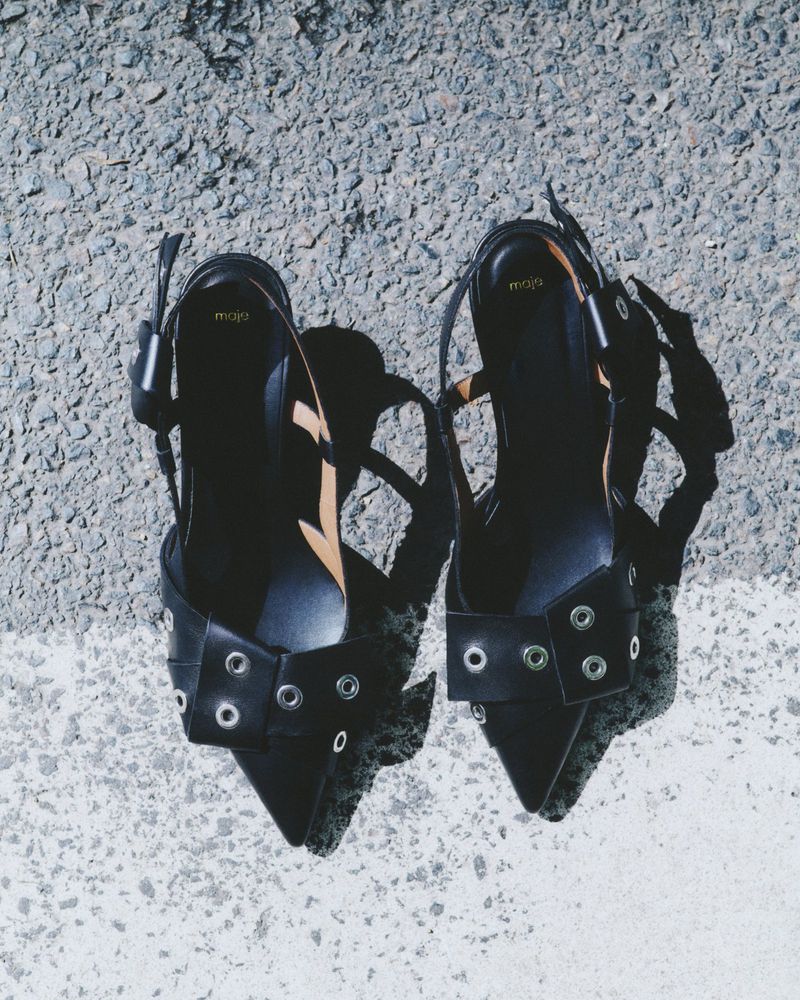 Two black Maje slingback shoes with metallic ring accents sit on a grey concrete surface with a bright white painted stripe.