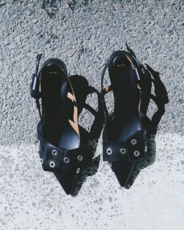 Two black Maje slingback shoes with metallic ring accents sit on a grey concrete surface with a bright white painted stripe.