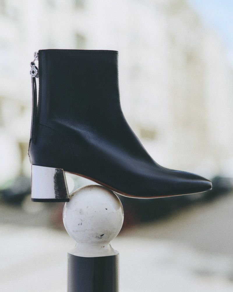 Elegant black ankle boot with a silver heel perched atop a white bollard against a blurred urban backdrop, fashion-forward footwear.