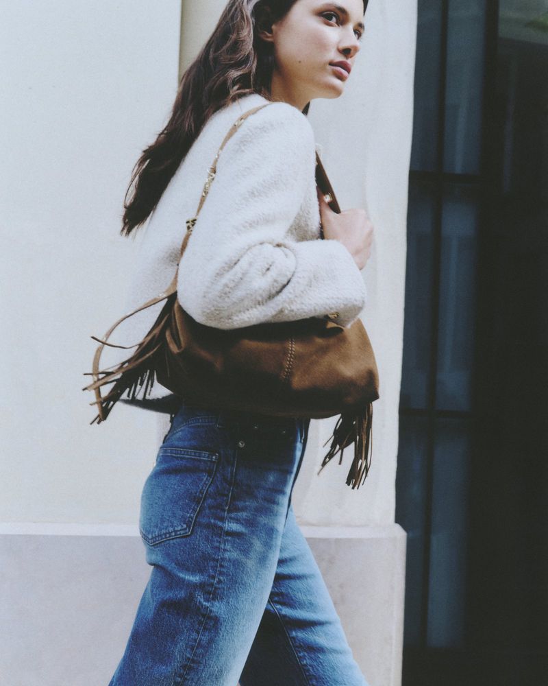 A woman walks outdoors carrying a brown fringe purse with a white jacket and jeans, capturing a candid fashion moment in natural light.