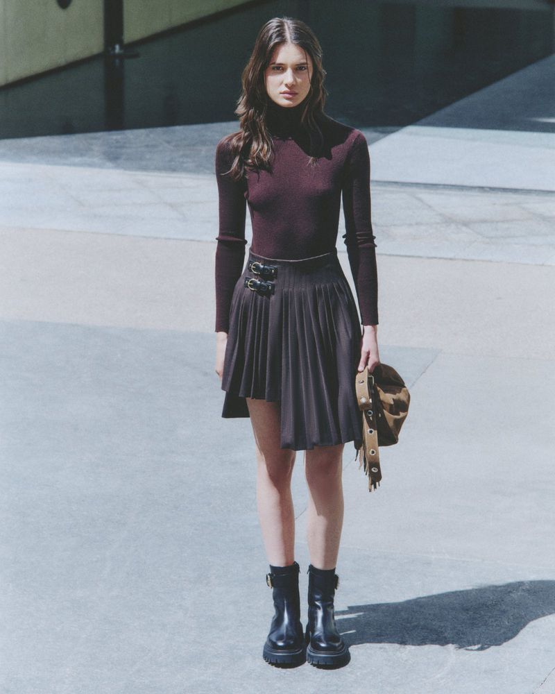 Model wearing a dark turtleneck and pleated skirt holding a handbag on an outdoor paved street for Maje FW24 Lookbook
