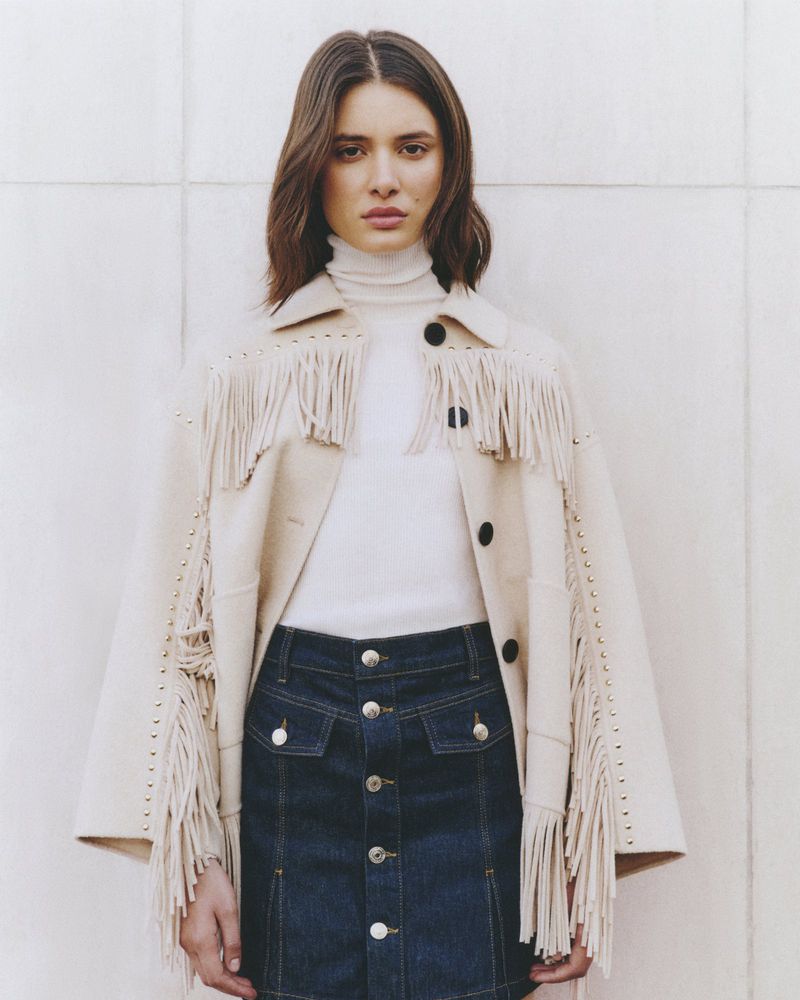 Woman wearing fringed jacket and denim skirt poses against a cream wall, showcasing a fashion-forward, contemporary Western style.