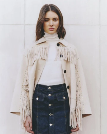 Woman wearing fringed jacket and denim skirt poses against a cream wall, showcasing a fashion-forward, contemporary Western style.