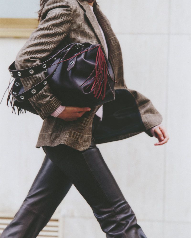 Fashionable woman strides forward holding a black leather bucket bag with tassels, wearing a plaid blazer and leather pants on city street.