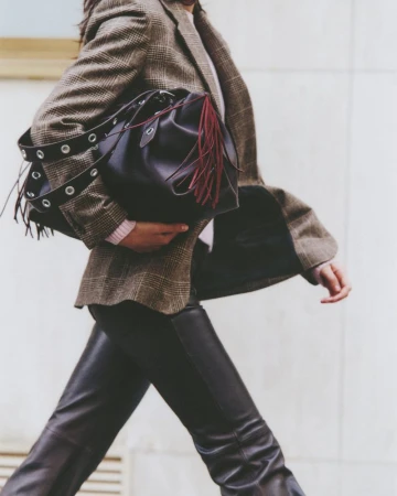 Fashionable woman strides forward holding a black leather bucket bag with tassels, wearing a plaid blazer and leather pants on city street.