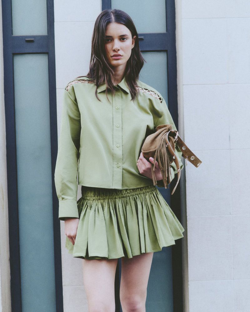 Woman wearing a light green, long-sleeved button-up shirt and a matching mini skirt poses against a light-colored building with windows.