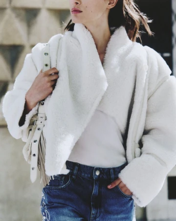 A stylish adult female wearing a white faux fur jacket, jeans, and a fringe bag in an urban environment. Fashion editorial photography.