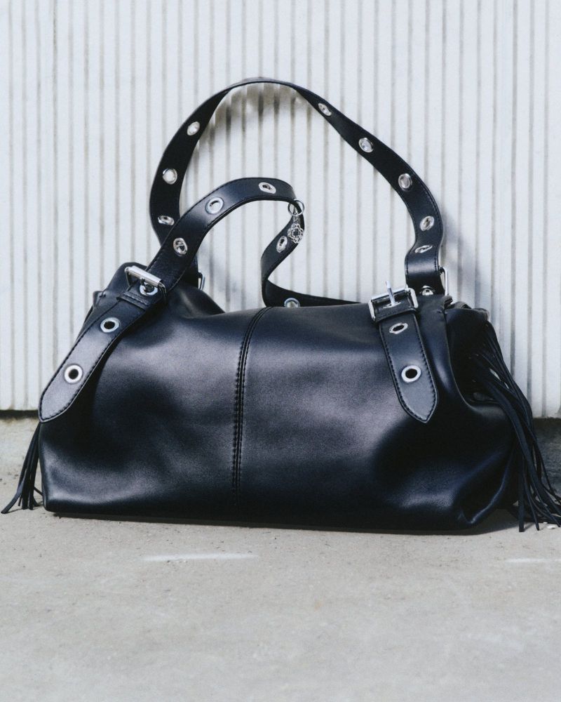 Studio shot of a black leather satchel bag with fringe detail and silver hardware against a vertically paneled wall and concrete floor.
