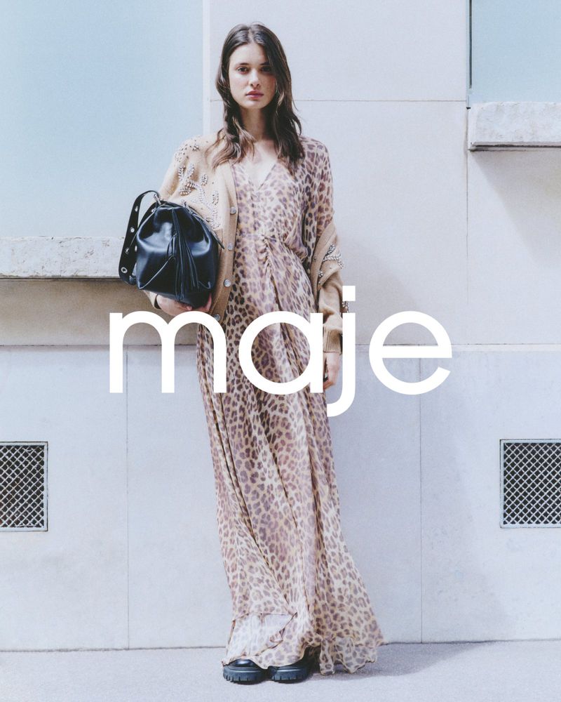 Fashion shot of a woman wearing an animal print maxi dress and a tan cardigan, holding a black bucket bag against a white wall.