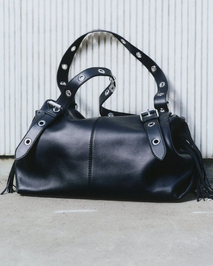 Black leather shoulder bag with metal grommets and fringe details resting against a white paneled wall