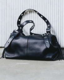 Black leather shoulder bag with metal grommets and fringe details resting against a white paneled wall