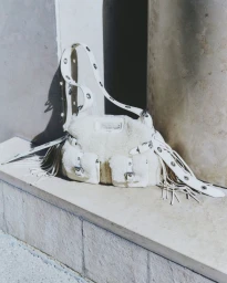 White shearling handbag with fringe and buckles resting on a stone ledge outdoors.