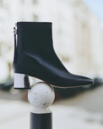 Black leather ankle boot with metallic silver block heel balanced delicately atop a white bollard sphere outdoors